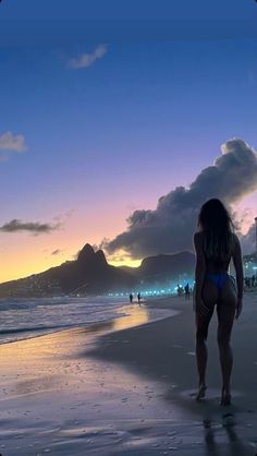 a woman standing on top of a sandy beach next to the ocean at sunset with mountains in the background