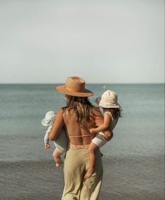 a woman walking on the beach with her baby in her arms and wearing a hat