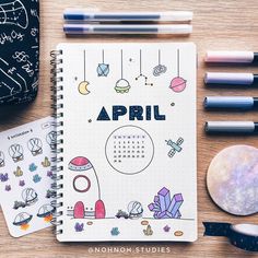 an open spiral notebook sitting on top of a wooden table next to markers and pens