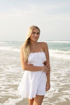 a beautiful young woman standing on top of a sandy beach next to the ocean wearing a white dress