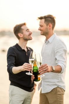 two men standing next to each other holding wine glasses in front of the ocean at sunset