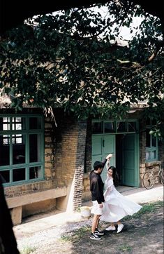 two people standing in front of a building with green doors and windows, one person reaching up into the air