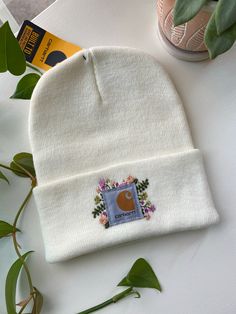 a white beanie with the letter c embroidered on it next to some flowers and a plant