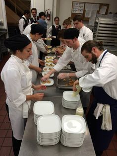 several chefs preparing food in a kitchen with plates and bowls on the counter, while others look on