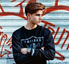 a young man standing in front of a graffiti covered wall with his arms crossed and looking at the camera