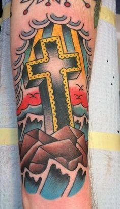 a cross tattoo on the arm with mountains and clouds in the background, done by person