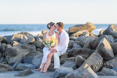 a bride and groom sitting on rocks at the beach