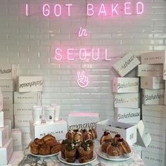 a table topped with lots of pastries on top of white boxes next to a neon sign that says i got baked in seoul