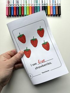 someone holding up a coloring book with strawberries on it and crayons in the background