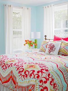 a bed with colorful comforters and pillows in front of two large windows that have white drapes on them