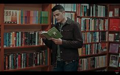 a man standing in front of a bookshelf holding a book and looking at it