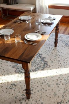 a wooden table with plates and glasses on it in front of a window next to a rug