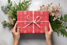 two hands holding a red gift box with a pink ribbon and bow on it, surrounded by flowers