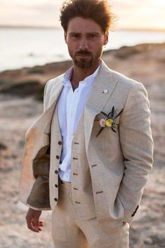 a man in a suit standing on the beach with his hands in his pockets and looking at the camera