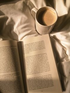 an open book sitting on top of a bed next to a cup of coffee