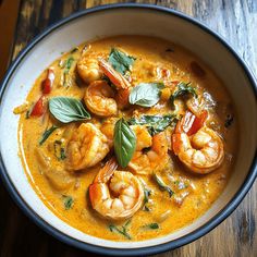 a bowl of soup with shrimp and basil garnishes on top, sitting on a wooden table