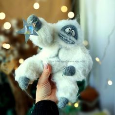 a hand holding a white stuffed animal in front of a christmas tree with lights on it