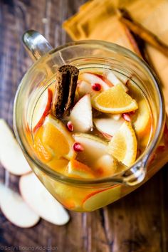 a glass mug filled with fruit and cinnamon on top of a wooden table next to sliced apples