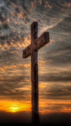 a wooden cross sitting on top of a hill under a cloudy sky with the sun setting behind it