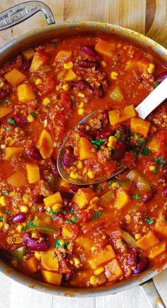 a large pot filled with chili and vegetables on top of a wooden table next to a spoon