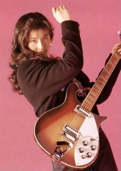 a woman holding an electric guitar in her right hand and posing for the camera with one arm up