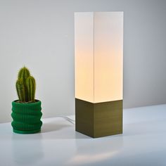 a cactus in a green pot next to a white lamp on a table with the light turned on