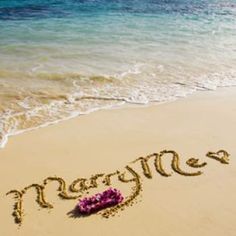 the word marry written in the sand at the beach with flowers on it's side