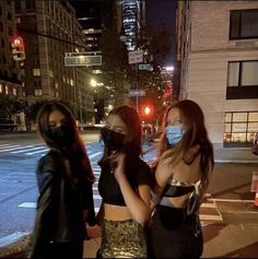 three women wearing face masks are standing on the street at night in front of a traffic light