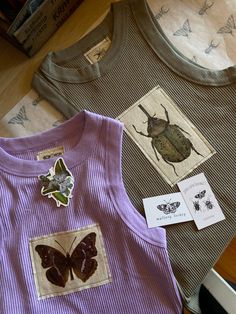 three t - shirts with bugs and butterflies on them sitting on a table next to some books