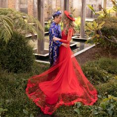 a man and woman dressed in red standing next to each other near some bushes with flowers