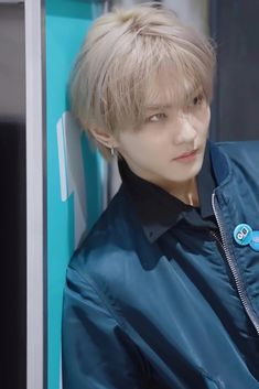 a young man with blonde hair wearing a blue jacket and earrings, leaning against a wall