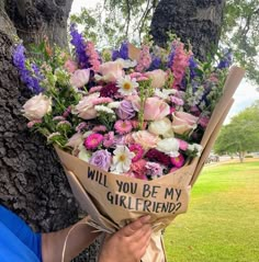 a woman holding a bouquet of flowers in front of a tree that says will you be my girlfriend?