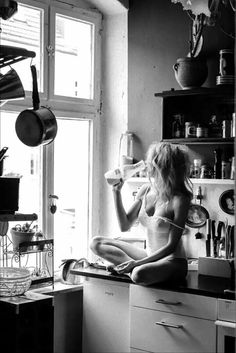 a woman sitting on top of a kitchen counter drinking from a cup in front of a window