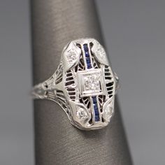 This lovely dinner ring is graceful and elegant, highlighting both diamonds and sapphires. The center diamond is an Old European cut stone measuring about 2.7mm (approx. 0.07ct). The stone is bright white G/H color and VS clarity. The ring also features four corner round Old European cut diamonds measuring 2mm (0.12cttw). In a stately column there are eight french cut blue sapphires highlighting the rest of the ring. The ring also has engraved shoulders. It is crafted in 18k white gold (stamped) Dinner Ring, French Cut, European Cut Diamonds, Antique Art Deco, G H, Cocktail Ring, Antique Art, Cocktail Rings, Bright White