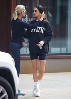 two women in matching sweatshirts talking to each other on the sidewalk near a car