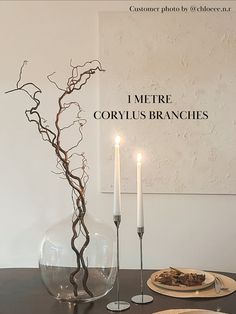 a table topped with plates and candles next to a glass vase filled with branches on top of a wooden table