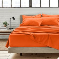 a bed with orange sheets and pillows in front of a brick wall next to a window