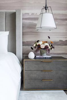 a nightstand with flowers on it in front of a bed and wallpapered headboard