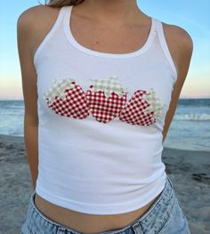 a woman standing on the beach wearing a white tank top with red and white checkered heart applique