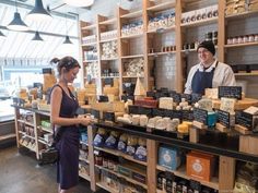 two people standing in front of a counter with cheeses and other items on it