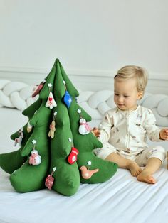 a baby sitting on a bed next to a green christmas tree pillow that is shaped like a toy