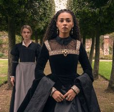 two women in black dresses standing next to each other on the grass and some trees