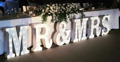 an illuminated mr and mrs sign with flowers on the table in front of it at a wedding reception