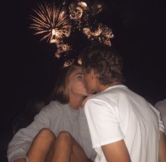 a man and woman sitting next to each other with fireworks in the sky behind them