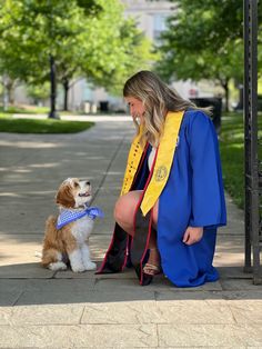 a woman in graduation gown kneeling down next to a dog on the sidewalk with trees behind her