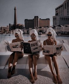 two women sitting on the edge of a swimming pool while wearing towels and reading newspapers