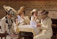 three women sitting at a table with hats on and papers in front of their faces