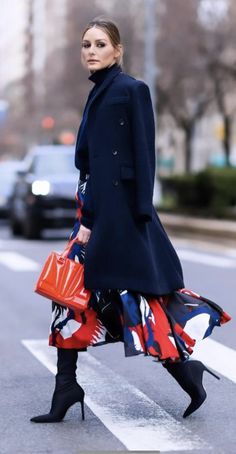 a woman is walking down the street wearing black boots and a blue coat with an orange bag