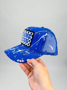 Score style points with this Game Day Checkered Splatter Paint Cap! The unique splatter design and checkered patch add a fun twist to a classic trucker hat. Perfect for showing your support for the blue team on game day. Key Features Adjustable snapback Lightweight + Comfortable Checkered Game Day Patch Splatter paint Pearl details Trendy Blue 5-panel Baseball Cap, Blue Snapback Hat With Letter Print And Curved Bill, Blue Curved Bill Baseball Cap For Game Day, Blue Sports Fan Snapback Hat For Game Day, Blue Snapback Hat For Game Day, Blue Snapback Hat For Sports Fans On Game Day, Blue Trucker Hat With Letter Print And Curved Visor, Blue 5-panel Trucker Hat For Baseball Season, Blue Letter Print Snapback Hat For Streetwear