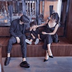 three people sitting on a bench in front of a store window, one woman is combing the mans hair